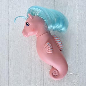May include: A pink seahorse toy with a smooth, matte finish, featuring a long, flowing mane of bright turquoise hair. The figure has large, dark purple eyes, a curved tail, and small, moulded fins along its back and side with light blue accents.