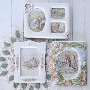 Könnte beinhalten: Drei weiße Keramik-Bilderrahmen mit floralen Akzenten und Illustrationen von Tieren und Menschen. Die Rahmen sind rechteckig und oval, mit einem Mehrfach-Fotorahmen oben. Auf einer weißen Holzoberfläche.
