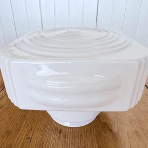 May include: A white, geometric ceiling light fitting with a square shape and a circular design on top. The fitting has a glossy finish and is mounted on a white base. The light fitting is made of glass.