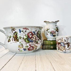 May include: A collection of antique white ceramic pieces with floral designs. The set includes a large chamber pot with a branch-shaped handle, a small vase, and a mug. The floral pattern features red, blue, and green hues.