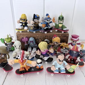 Puede incluir: Una colección de coloridas figuras de monopatín en miniatura. Las figuras presentan varios personajes con atuendos y accesorios únicos, cada uno montado en un monopatín. La colección incluye una variedad de colores y diseños, exhibidos sobre una superficie de madera blanca.