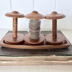 May include: Three brown wooden spools arranged on a matching wooden tray. Two spools are empty, while the centre spool is wrapped with a thick, textured grey yarn. The spools feature a classic cylindrical design with flat circular ends.