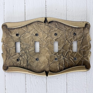 Könnte beinhalten: Dekorative Lichtschalterabdeckung mit vier Kippschalteröffnungen. Die Abdeckung hat ein strukturiertes, zerknittertes Papieraussehen in Gold- und Brauntönen, umrahmt von einem goldfarbenen Rand. Das Design hat eine Vintage- und Antik-Ästhetik.