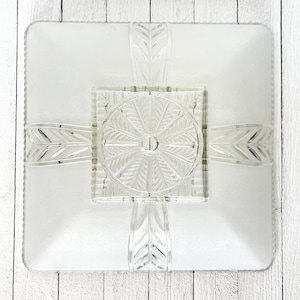 May include: A clear glass ceiling light fitting with a square shape and a geometric design. The light fitting has a central circular opening and four arms that extend out to the corners of the square.