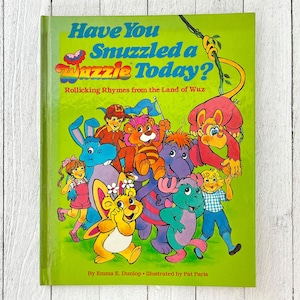 Op de afbeelding: Een groene boekomslag met de titel "Have You Snuzzled a Wuz zle Today?" in kleurrijke letters. Het boek bevat illustraties van cartoonfiguren, waaronder een paarse olifant, een gele beer, een blauw konijn en een groene kikker. Het boek is getiteld "Rollicking Rhymes from the Land of Wuz" en is geschreven door Emma E. Dunlop, geïllustreerd door Pat Paris.