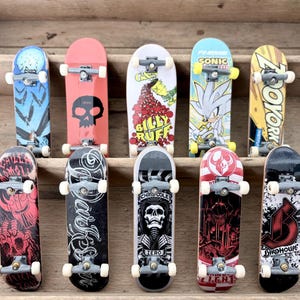 Puede incluir: Una colección de diez monopatines en miniatura con varios diseños. Las tablas presentan gráficos que incluyen calaveras, patrones abstractos y personajes como Sonic the Hedgehog. Cada tabla tiene cuatro ruedas blancas y ejes plateados.