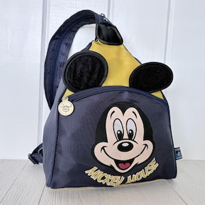 May include: A navy blue and yellow sling backpack featuring a graphic of Mickey Mouse's face and the text "MICKEY MOUSE" printed below. The bag includes two black circular ears attached to the top, a single shoulder strap, and a gold zipper pull.