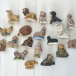 Peut inclure: Une collection de petites figurines en céramique représentant divers animaux et personnages, dont un papillon, un lion, un hérisson, un phoque, un chien, un ours, une personne en cape à capuche et d'autres créatures dans des tons marron, beige et blanc.