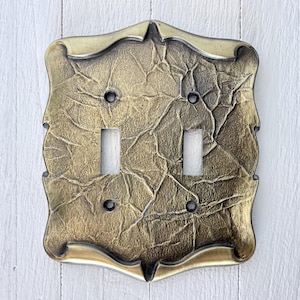 May include: A decorative double light switch cover with a textured, antique gold finish. The cover has a detailed, ornate design with two toggle switch openings and four screw holes. The edges are slightly curved and have a lighter gold trim.
