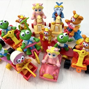May include: A collection of colourful Muppet Show character toys. Kermit the Frog, Fozzie Bear, and other characters are featured on various vehicles, including a pink car, red wagons, and a yellow tricycle. The toys are made of plastic and are set against a white background.