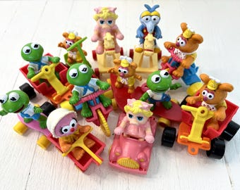 May include: A collection of colorful Muppet Show character toys. Kermit the Frog, Fozzie Bear, and other characters are featured on various vehicles, including a pink car, red wagons, and a yellow tricycle. The toys are made of plastic and are set against a white background.