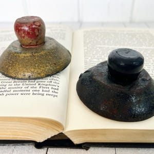 Può includere: Due ferma libri vintage in metallo con cime in legno. I ferma libri sono di colori diversi, uno è dorato con una cima rossa e l'altro è nero con una cima nera.