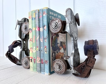May include: Vintage roller skate bookends in silver metal, holding a stack of colourful children's books. The bookends have brown leather straps and blue felt padding. The books have illustrations and the text "Best in Children's Books".