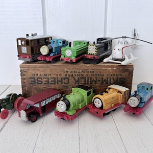 May include: A selection of toy vehicles, including trains, a bus, a helicopter, and a tractor. The trains are in various colours, such as blue, green, yellow, and brown. The bus is red, and the helicopter is white. The toys are on a wooden box.