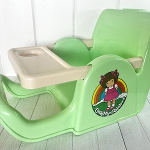 May include: A vintage green plastic baby doll high chair with a removable tray. The chair features a "Little Miss Rainbow" decal with a cartoon girl and rainbow. The chair has a curved base and armrests, and a backrest for support.
