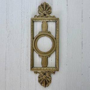 May include: A vintage brass wall sconce with a rectangular frame and a circular opening. The sconce features ornate floral and shell-like details at the top and bottom. The brass has a warm, aged patina. The sconce is mounted on a white wooden surface.