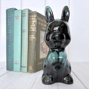 May include: A glossy, dark green and black ceramic rabbit figurine. The rabbit is sitting upright with long ears and a detailed face. Behind the rabbit are four vintage books with various colours and titles.