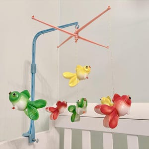 May include: A cot mobile featuring a light blue plastic support arm and a pink cross-shaped hanger. Five colourful goldfish figures in shades of green, yellow, and red are suspended from the hanger by thin white strings, creating a hanging display.