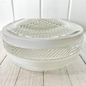 May include: A clear glass ceiling light fitting with a textured, square top and a white, rounded base. The glass has a patterned design. The fitting is on a white wooden surface.