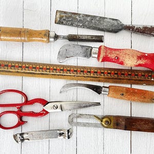 May include: A collection of vintage tools including a wooden ruler, a red and silver pair of scissors, a small wooden handled knife, a metal chisel, a metal and wooden handled utility knife, and a metal and wooden handled scraper.
