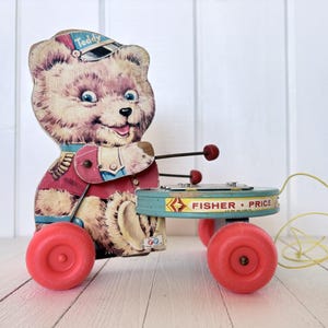 Könnte beinhalten: Vintage Fisher-Price Teddybär Xylophon-Spielzeug. Das Spielzeug zeigt einen lächelnden Bären in roter und weißer Uniform mit blauem Hut. Das Xylophon ist hellblau mit roten Rädern und einer gelben Schnur. Der Text "Fisher-Price" ist sichtbar.