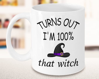 May include: A white ceramic mug featuring the black printed text "TURNS OUT I'M 100%" above a graphic of a black witch hat with a purple band and gold buckle, followed by the black printed text "that witch" centered on the front surface.