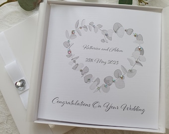 May include: A white card with a silver eucalyptus wreath in the shape of a heart. The card says "Katherine and Adam 28th May 2023" and "Congratulations on your wedding".