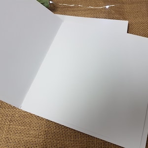May include: Blank white card with a folded design. The card is on a brown burlap surface.