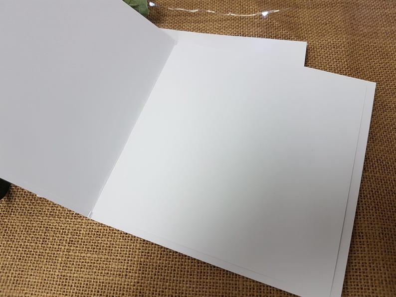 May include: Blank white card with a folded design. The card is on a brown burlap surface.