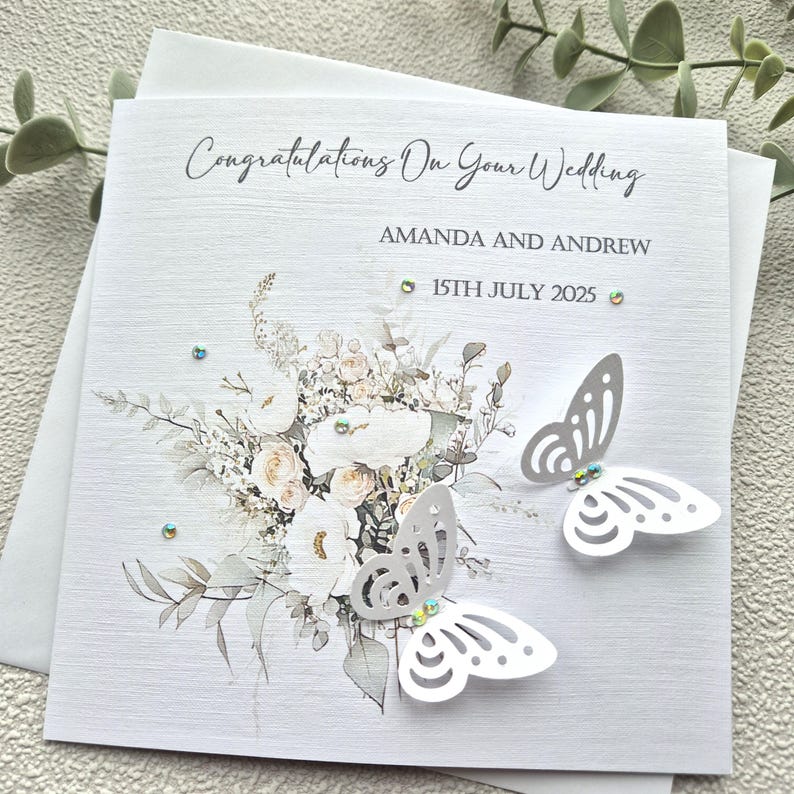 May include: A white wedding card with a floral design and two white butterflies. The card reads "Congratulations on your wedding" and "Amanda and Andrew" with the date "15th July 2025".