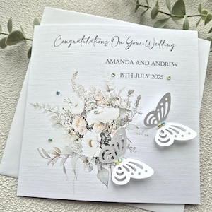 May include: A white wedding card with a watercolor floral design and two white paper butterflies. The card reads "Congratulations on your wedding" and includes the names "Amanda and Andrew" and the date "15th July 2025".