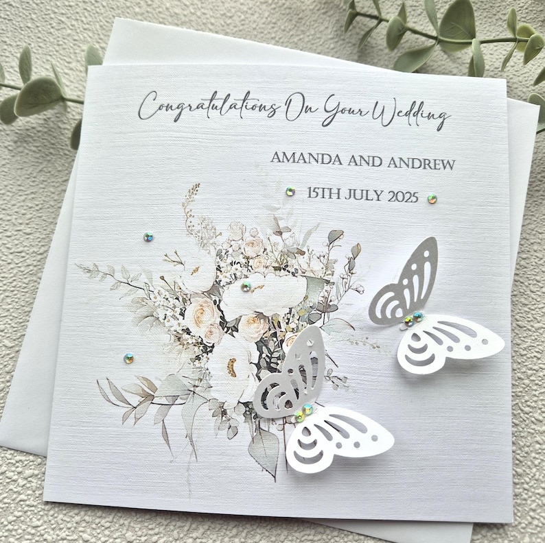 May include: A white wedding card with a watercolor floral design and two white paper butterflies. The card reads "Congratulations on your wedding" and includes the names "Amanda and Andrew" and the date "15th July 2025".