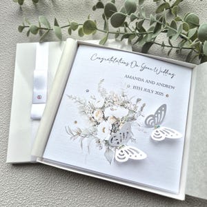 May include: A white wedding card with a floral design and two white paper butterflies. The card reads "Congratulations on your wedding Amanda and Andrew 15th July 2025".