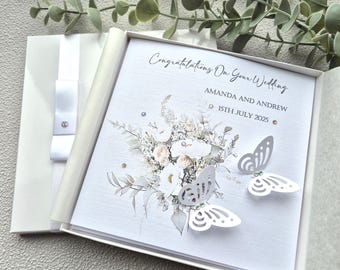 May include: A white wedding card with a floral design and two white paper butterflies. The card reads "Congratulations on your wedding Amanda and Andrew 15th July 2025".