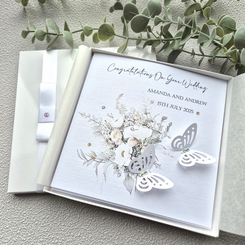 May include: A white wedding card with a floral design and two white paper butterflies. The card reads "Congratulations on your wedding Amanda and Andrew 15th July 2025".