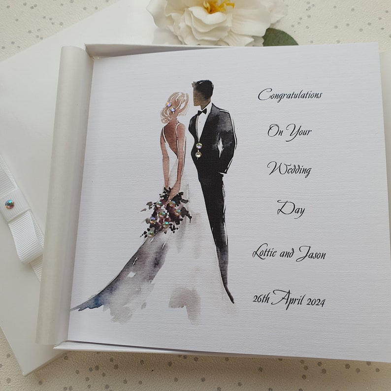 May include: A white card with a watercolor illustration of a bride and groom. The text reads "Congratulations on your Wedding Day Lottie and Jason 26th April 2024".