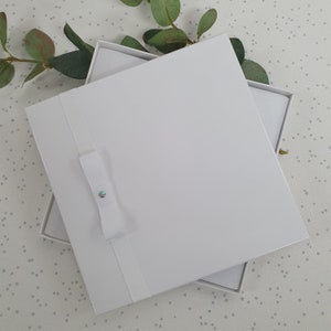 May include: White gift box with a white ribbon and a small, iridescent rhinestone. The box is open and the inside is also white.