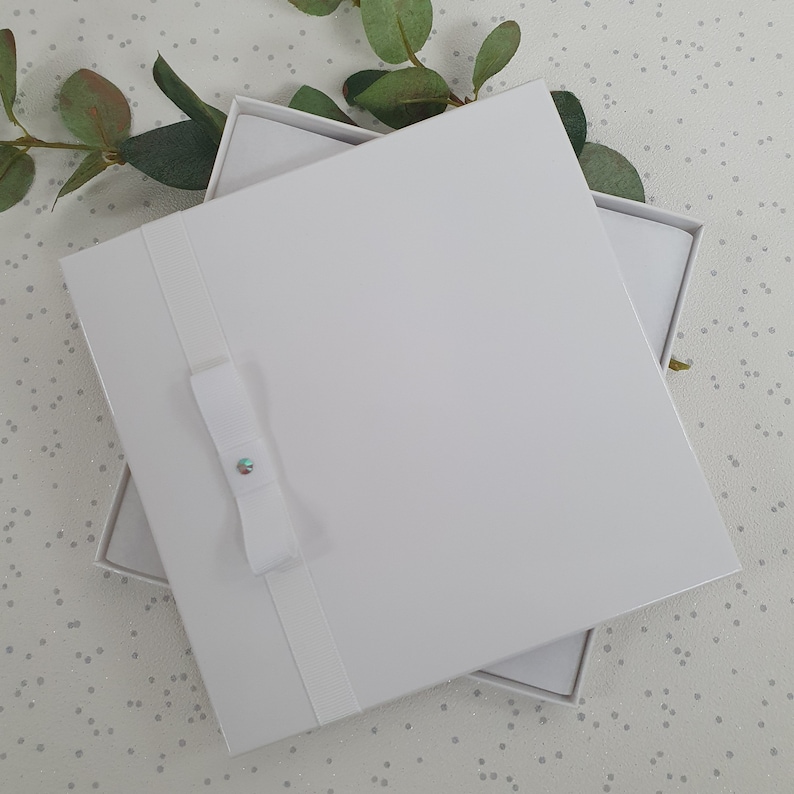 May include: White gift box with a white ribbon and a small, iridescent rhinestone. The box is open and the inside is also white.