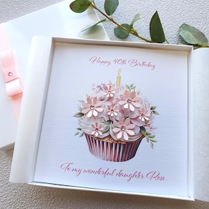 May include: A birthday card in a white box with a pink ribbon. The card shows a cupcake design with pink flowers and a lit candle. Text reads "Happy 40th Birthday" and "To my wonderful daughter Rose."