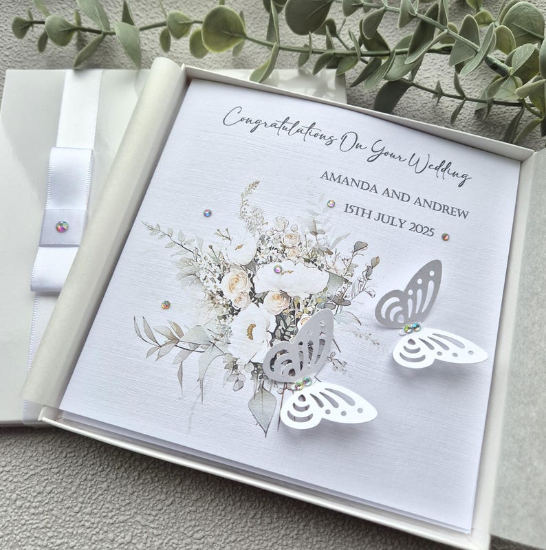 May include: White wedding card with a floral design and two white butterflies. The card reads "Congratulations On Your Wedding" and "Amanda and Andrew 15th July 2025".