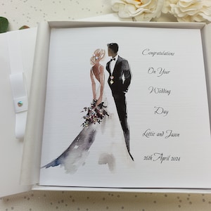 May include: A white card with a watercolor illustration of a bride and groom. The text reads "Congratulations on your Wedding Day Lottie and Jason 26th April 2024".