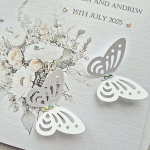 May include: A white wedding invitation with a floral design and two white paper butterflies with silver accents. The text on the invitation reads "[Name] and Andrew 15th July 2025".