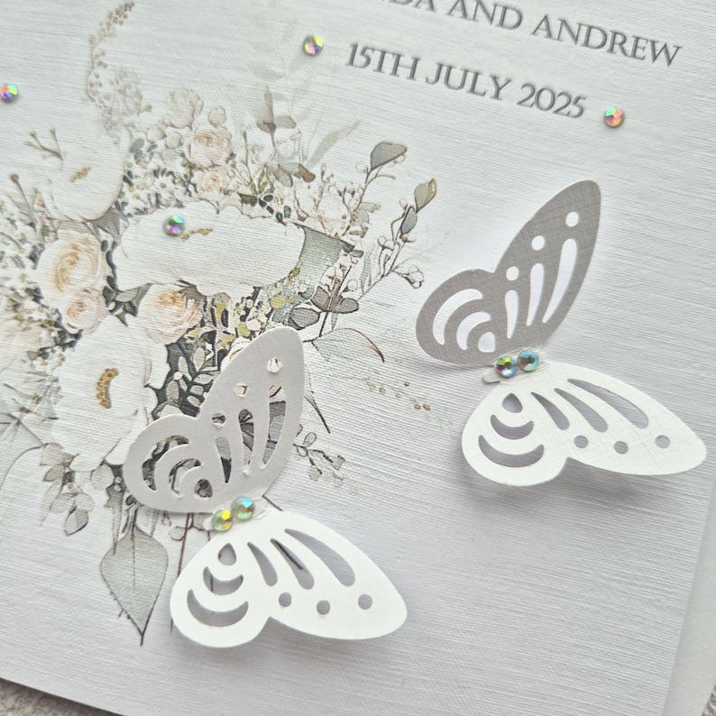 May include: A white wedding invitation with a floral design and two white paper butterflies with silver accents. The text on the invitation reads "[Name] and Andrew 15th July 2025".