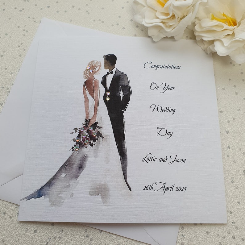 May include: A white wedding card with a watercolor illustration of a bride and groom. The text reads "Congratulations on your Wedding Day Lottie and Jason 26th April 2024".