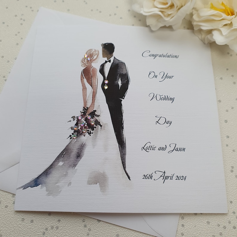 May include: A white wedding card with a watercolor illustration of a bride and groom. The card reads "Congratulations on your Wedding Day Lottie and Jason 26th April 2024".