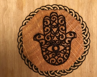Può includere: Un disegno di mano di hamsa bruciato sul legno con un occhio al centro, circondato da un bordo di nodo celtico.