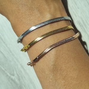 Puede incluir: Tres pulseras de cadena de serpiente plateadas, doradas y de oro rosa apiladas en la muñeca de una persona.