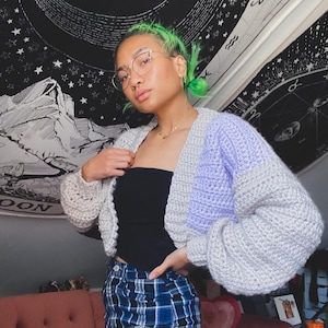 May include: A woman with green hair is wearing a light grey and purple crocheted cardigan with a black tank top and blue and black plaid trousers.