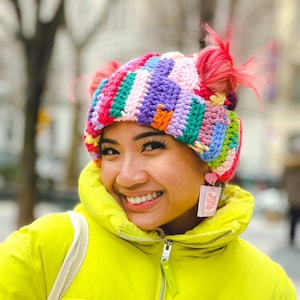 May include: A colorful, crocheted hat with pink accents and two fluffy pink pom-poms. The hat features a patchwork design with various colors. The person is wearing a bright yellow puffer jacket with the word "FOLLOWERS" on the front.