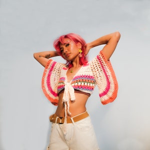 May include: A woman with pink hair wearing a white crochet top with pink, orange, and green stripes. The top has a tie at the front and is cropped. She is also wearing a white pair of pants with a brown belt.